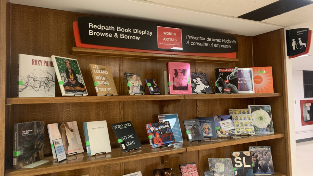 Brown bookshelf with colourful books. On the top, there is a red sign that says Women Artists. 