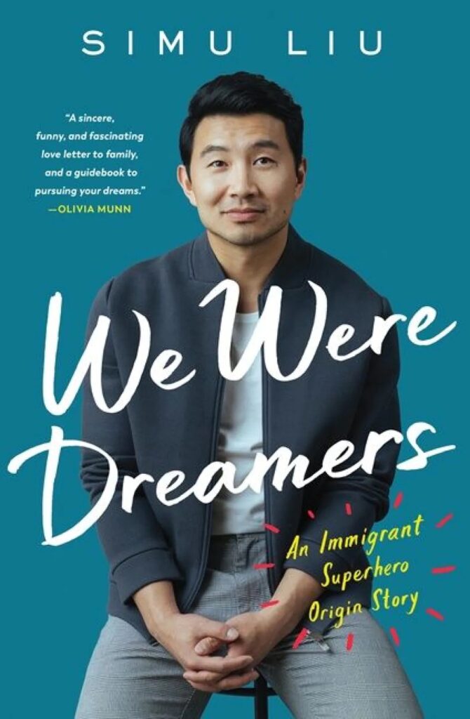 Book Cover for We Were Dreamers by Simu Liu. The cover features Simu Liu sitting on a stool. He is a handsome Asian-Canadian man wearing light grey pants, a white shirt, and navy/dark grey jacket. His hands are crossed in front of his lap.