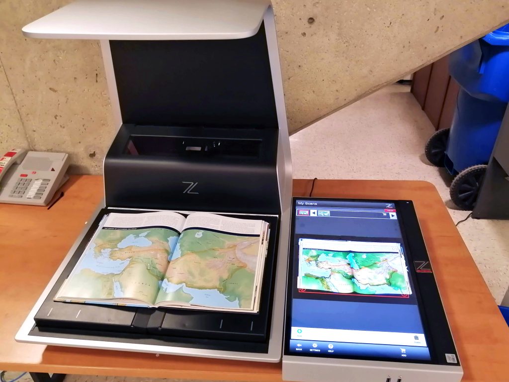 The Zeta overhead scanner is a large machine sitting on a desk with an open book sitting on top of the scanner. Beside the machine is a digital image of the book being scanned.