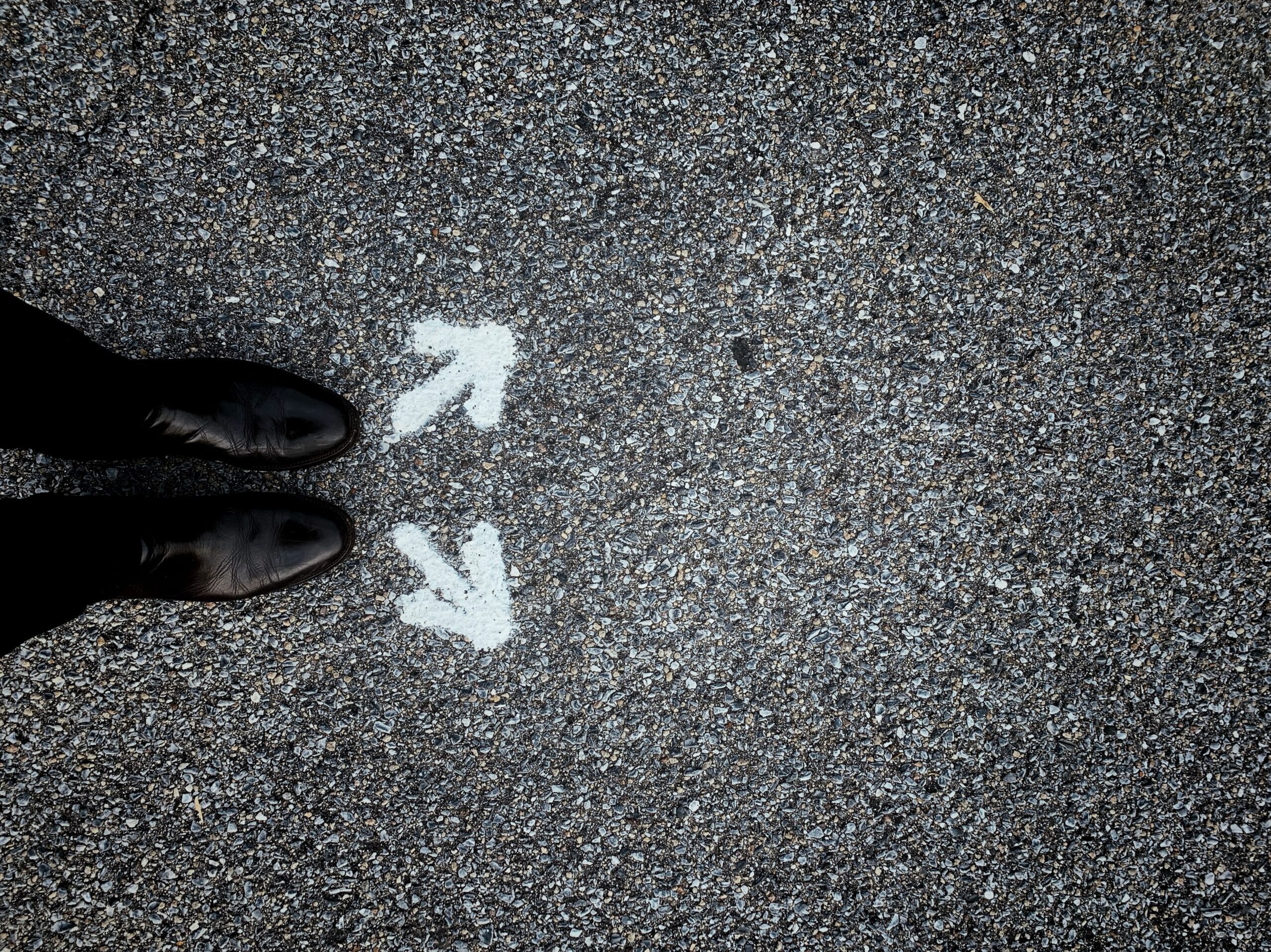Picture from above of black shoes and bottom of black pants standing on black asphalt, with two white arrows painted on the asphalt just above the feet, one arrow pointing diagonally up and the other pointing diagonally down